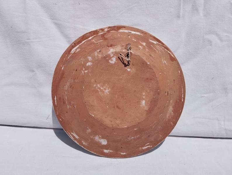 Op de afbeelding: Een ronde terracotta schaal met een roodbruine afwerking en zichtbare witte verfresten op het oppervlak. De achterkant van de schaal is voorzien van een klein metalen draadlusje nabij het midden, bedoeld om het object aan op te hangen.