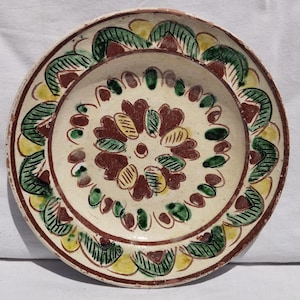 Op de afbeelding: Een ronde keramische bord met een cr&egrave;mekleurige basis, voorzien van handgeschilderde bloem- en bladmotieven in tinten donkerrood, bosgroen en mosterdgeel. Het ontwerp bevat een centraal bloempatroon omringd door een decoratieve rand.