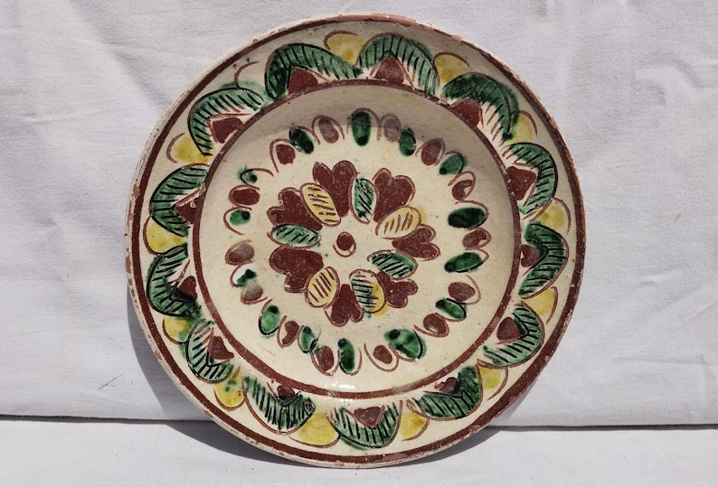 Op de afbeelding: Een ronde keramische bord met een cr&egrave;mekleurige basis, voorzien van handgeschilderde bloem- en bladmotieven in tinten donkerrood, bosgroen en mosterdgeel. Het ontwerp bevat een centraal bloempatroon omringd door een decoratieve rand.