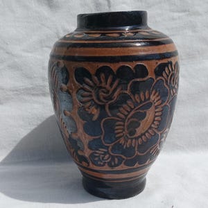 Può includere: Un vaso di ceramica con un motivo floreale blu scuro e marrone. Il vaso ha un corpo arrotondato e un collo stretto. Il motivo floreale è scolpito sulla superficie, creando un effetto strutturato. Il vaso è alto circa 20 cm.