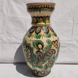 Può includere: Un vaso in ceramica con base color crema, ornato con motivi floreali verdi, gialli e marroni. Il vaso ha una parte superiore ampia e svasata e un corpo bulboso, che mostra un design tradizionale.
