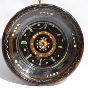 Può includere: Un piatto in ceramica marrone con un design geometrico in bianco e nero. Il piatto ha un bordo marrone con un motivo ondulato e un centro bianco con un cerchio di punti verdi e marroni.