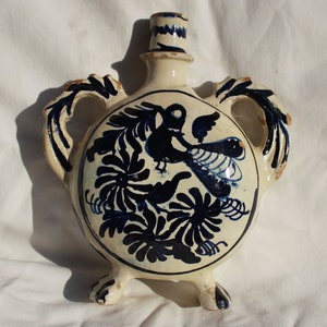 May include: A white ceramic bottle with blue and black floral designs and a bird. The bottle has two handles and three legs.