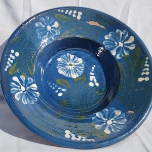 Könnte beinhalten: Ein handbemalter, blauer Keramikteller mit floralem Design. Der Teller zeigt weiße und blaue Blumen mit grünen Blättern. Der Rand und die Mitte des Tellers sind dunkler blau.