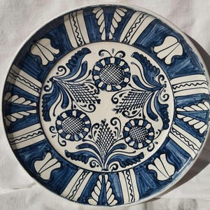 Op de afbeelding: Een ronde keramische plaat met een witte basis en ingewikkelde blauwe bloemmotieven. De plaat heeft een centraal bloemmotief, omgeven door gestileerde bladeren en een patroonrand. De diameter van de plaat is ongeveer 25 cm.