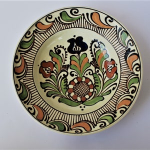 May include: A white ceramic plate with a hand-painted floral design. The design features green, orange, and black flowers with a brown centre. The plate has a black and white border.