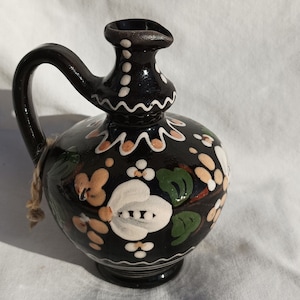 May include: A black ceramic pitcher with a handle and spout, adorned with white, peach, and green floral designs. The pitcher has a decorative rim and a natural fiber string tied around the neck. The background is a white cloth.