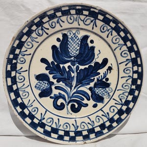 May include: A round, antique ceramic plate with a white base and blue floral design. The plate's border features a checkered pattern and decorative swirls. The central floral motif is a stylized blue flower with detailed leaves.