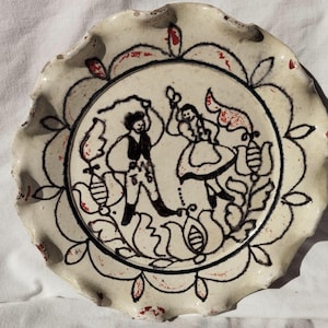Può includere: Piatto in ceramica bianco sporco con bordo smerlato e design nero e rosso. L'immagine centrale raffigura due figure che ballano, circondate da motivi floreali e di uccelli. Il piatto ha un aspetto rustico e fatto a mano.