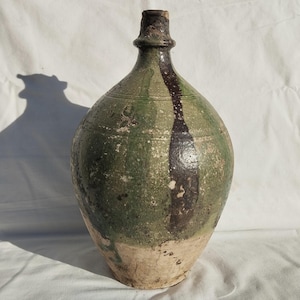 Op de afbeelding: Een grote, antieke keramische kruik met een bolvormige body en een smalle hals. De kruik is voornamelijk groen met een donkerbruine verticale streep en een lichtbeige basis. De hoogte van de kruik is ongeveer 46 cm.