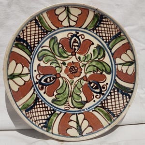 May include: A round, hand-painted ceramic plate with a cream-coloured base. The plate features a central floral design in shades of red, green, and brown, surrounded by a blue and black geometric pattern. The plate's diameter is approximately 25 cm.