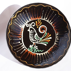 Può includere: Piatto rotondo in ceramica nera con un motivo decorativo di uccello al centro. L'uccello è bianco con accenti colorati di verde, arancione e giallo. Il bordo ha un bordo smerlato con dettagli marroni e bianchi.