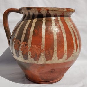 May include: A rustic, hand-painted ceramic jug with a handle. The jug features vertical stripes in shades of red, brown, and cream. The earthenware vessel is likely used for serving beverages or as a decorative item.