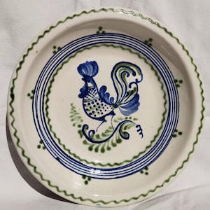 Puede incluir: Un plato de cerámica blanco con un diseño de gallo azul y verde. El plato presenta un borde festoneado con detalles verdes y círculos concéntricos azules. El gallo está en el centro, con detalles azules y verdes.