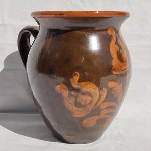 Puede incluir: Jarra de cerámica marrón con asa y diseño floral naranja. La jarra tiene un acabado brillante y un borde naranja. Esta pieza de cerámica artesanal es adecuada para servir bebidas, con una capacidad de aproximadamente 1,5 litros.