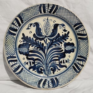May include: A round ceramic plate featuring a cream-coloured base with hand-painted dark blue floral motifs in the centre. The rim is decorated with a repeating pattern of blue cross-hatching and vertical stripes, creating a symmetrical design.