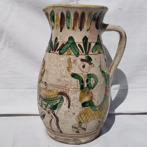 Puede incluir: Jarra de cerámica vintage con asa, con figuras y patrones pintados a mano en verde, amarillo, marrón y rosa. La jarra tiene una base de color crema y un aspecto ligeramente desgastado. Es un artículo decorativo.