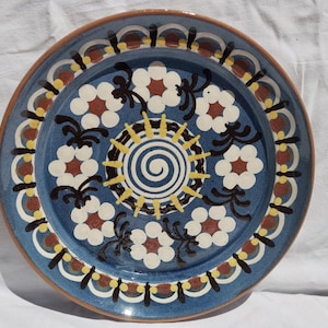 May include: A round ceramic plate featuring a central blue and white spiral design surrounded by a ring of white flowers with red centers. The plate has a blue background with black leaf accents and a decorative border of yellow and brown patterns.