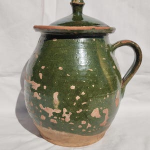 May include: A green ceramic jug with a lid and handle. The jug has a rounded body and a dark green glaze with lighter brown patches. The lid is also green with a small knob. The jug is likely vintage or antique.