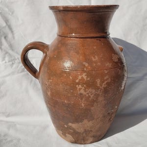 Può includere: Una brocca in ceramica marrone con manico e bocca larga. La brocca ha un corpo bulboso e una superficie strutturata con macchie più chiare. La brocca viene probabilmente utilizzata per conservare liquidi e ha un aspetto rustico e antico.