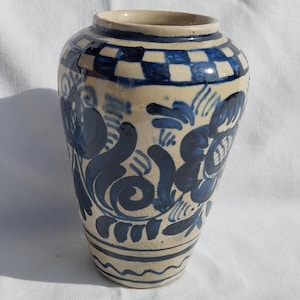 May include: A ceramic vase with a cream base and blue floral and geometric patterns. The vase has a wide body and a slightly flared rim. The blue design includes a checkered pattern at the top and floral motifs around the body.