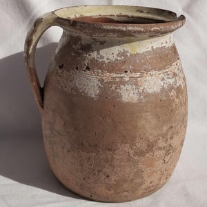 May include: A rustic, brown ceramic pitcher with a handle. The pitcher has a wide body and a narrow neck, with a cream-colored rim and handle. The paint is chipped, giving it a weathered appearance. A vintage piece.