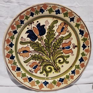Op de afbeelding: Een ronde, crèmekleurige keramische plaat met een bloemenontwerp. Het midden heeft een boeket van blauwe en oranje bloemen met groene bladeren. De rand is versierd met een herhalend patroon van blauwe, oranje en groene bloemmotieven.