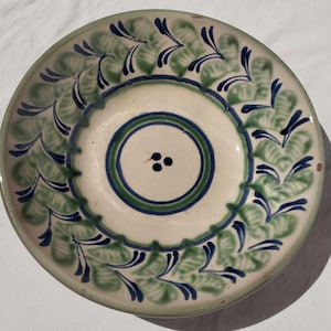 May include: A round ceramic plate with a cream-coloured base featuring a hand-painted pattern of green leaves and dark blue accents. The centre of the plate displays three small dark blue dots surrounded by concentric green and blue circular rings.