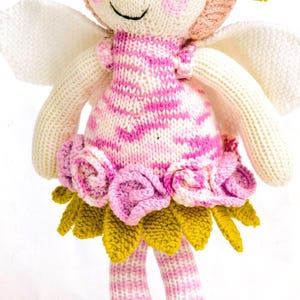 May include: A knitted fairy doll with a smiling face, rosy cheeks, and black eyes. The doll wears a pink and white striped dress, a flower crown, and has white wings. The dress is decorated with pink roses and yellow leaves.