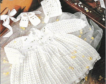 May include: A white crocheted baby dress with a matching hat and booties. The dress has a lace trim and a bow at the neck. The set is displayed on a white tulle fabric with white flower petals.