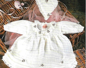 May include: A white knitted baby dress with a lace trim and floral embellishments. The dress is displayed in a wicker basket with a pink tulle fabric and a white knitted bonnet.