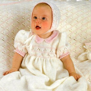 May include: A white knitted baby dress with a pink lace trim and a matching bonnet. The dress has a delicate lace pattern and is worn by a baby sitting on a white blanket.