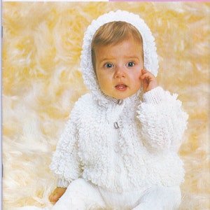 May include: A white knitted baby jacket with a hood and button closure. The jacket has a fluffy texture and is worn by a baby sitting on a fuzzy white surface.
