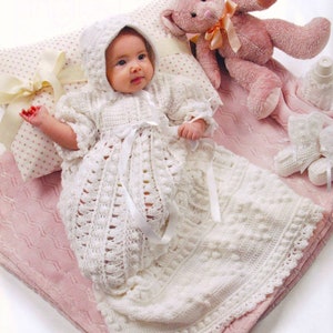 May include: A white crocheted baby gown with a white satin ribbon and a white crocheted bonnet. The gown is laid on a pink blanket with a teddy bear and other baby accessories.