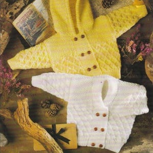 May include: Two knitted baby sweaters. One is yellow with a hood and brown buttons, the other is white. Both have a diamond pattern on the front. The sweaters are displayed on a rustic surface with a vintage map and decorative elements.