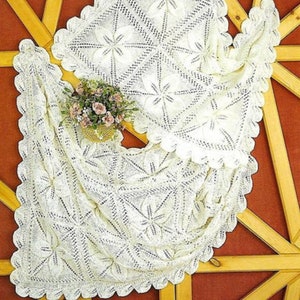 Puede incluir: Una manta de bebé de ganchillo blanco con un delicado patrón floral. La manta está doblada y se muestra sobre una superficie de madera.