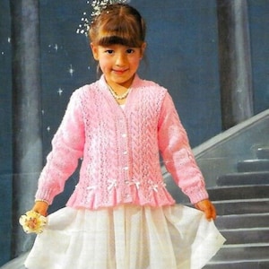 Puede incluir: Una niña pequeña lleva un cárdigan de punto rosa con ribete de encaje blanco y un collar de perlas. Lleva una falda blanca y zapatos blancos. Está de pie en un tramo de escaleras con flores en los escalones.