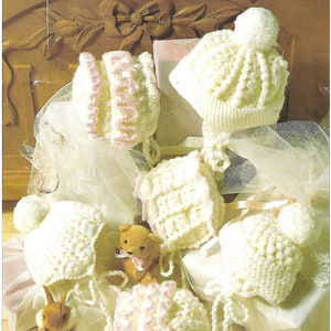May include: Five crocheted baby hats in white and pink with ties. The hats are arranged on a white fabric background with a teddy bear and a bunny rabbit. The text "PREMATURE CROCHET" is visible in the lower right corner.