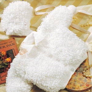 May include: A white knitted baby jacket and bonnet set with a looped texture. The jacket features a button closure. The set is displayed with a vintage book and pearl necklace, suggesting a classic, handmade baby clothing set.