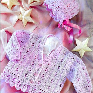 May include: A pink knitted baby cardigan and bonnet set with a delicate lace pattern. The set is laid out on a pink fabric background with decorative stars.
