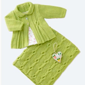 May include: A green knitted baby cardigan with a white collar and button closure, paired with a matching green knitted blanket with a cable knit pattern. A small heart-shaped ornament with floral embellishments is attached to the blanket.