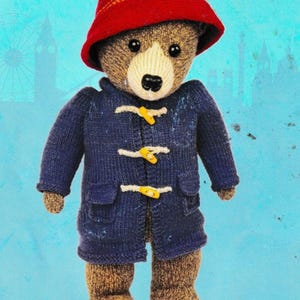 Könnte beinhalten: Gestrickte Paddington Bär Puppe, trägt einen roten Hut und einen marineblauen Mantel mit gelben Knebeln. Der Bär hat eine weiße Schnauze und braunes Fell. Eine charmante Darstellung des beliebten Kinderbuchcharakters.