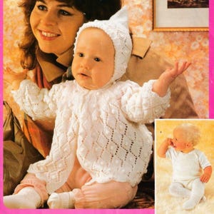 May include: A baby wearing a white knitted outfit, including a jacket and a hat. The jacket has a diamond pattern with small bobbles. The baby is also wearing pink pants and white socks. A woman is in the background.