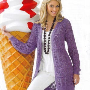 May include: A long, open-front, purple cardigan with a delicate, open-knit design. The cardigan features long sleeves and a button closure. The woman is wearing a white top and white pants. A large ice cream cone prop is in the background.