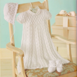 Pode incluir: Um vestido de bebé de malha branca com um chapéu e sapatinhos a condizer. O vestido tem um padrão de diamante e um decote festoneado. O conjunto é apresentado numa cadeira de madeira.