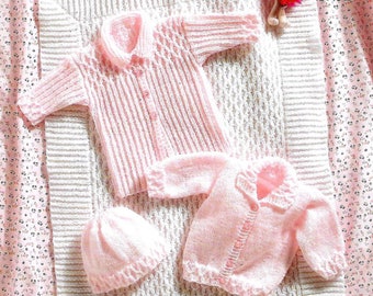 May include: A pink knitted baby outfit set including a cardigan, a shirt, and a hat. The set is laid out on a white crocheted blanket.