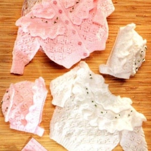 Puede incluir: Un conjunto de ropa de bebé en rosa pálido y blanco. El conjunto incluye un cárdigan, un gorro y patucos, todos con delicados detalles de encaje y florales. Los cárdigans tienen botones y bordes con volantes, ideal para un recién nacido.