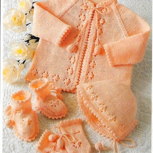 May include: A knitted baby outfit in a peach colour. The set includes a cardigan, a hat, booties, and mittens. The cardigan has a button fastening and a decorative trim. The hat has a scalloped edge. The booties and mittens have pom-poms.