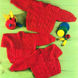 May include: Three red knitted sweaters and a set of toy kitchen utensils are displayed on a green surface. Two of the sweaters are cardigans with buttons, and one is a crew neck. The toys include a pan, plates, and measuring spoons.