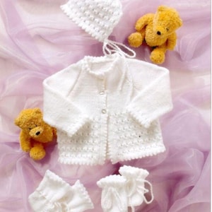 Puede incluir: Un conjunto de bebé blanco tejido, que incluye una chaqueta, un gorro y patucos, se muestra con dos pequeños osos de peluche amarillos. El conjunto presenta un patrón texturizado y cierres de botón. Los artículos están dispuestos sobre una tela suave de color lavanda.